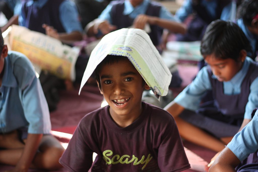 Intérieur, couleur, une salle de classe. Un jeune garçon portant un tee shirt violet est coiffé d'un journal. A ses côtés, les écoliers portent l'uniforme.