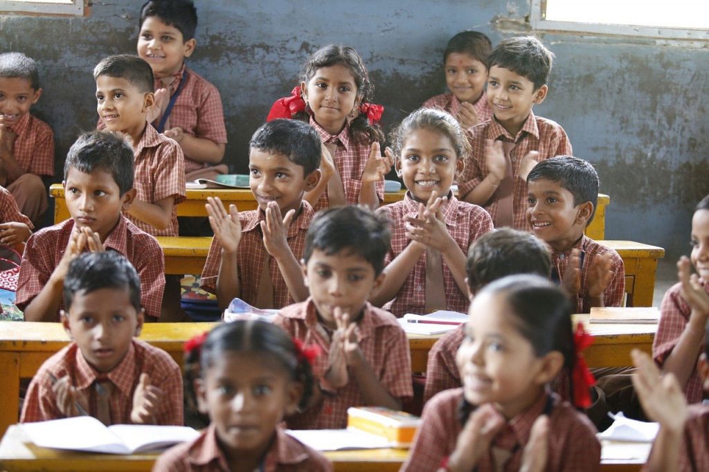 Une salle de classe, photo couleur. Des enfants en uniforme (chemisette à carreau et cravatte) écoutent le professeur en souriant. Certains frappent dans leurs mains. Sont-ils en train de mimer une comptine ?
