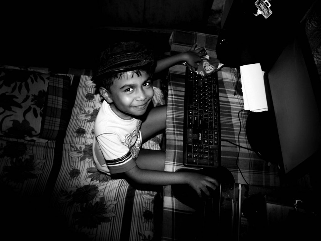 Interieur noir et blanc. Un très jeune garçon coiffé d'une caquette regarde avec malice et fierté le photographe. On aperçoit un clavier d'ordinateur posé sur une table devant lui. Sa main droite est posée sur la souris. Sa main gauche plonge dans un paquet de gateau apéritif.