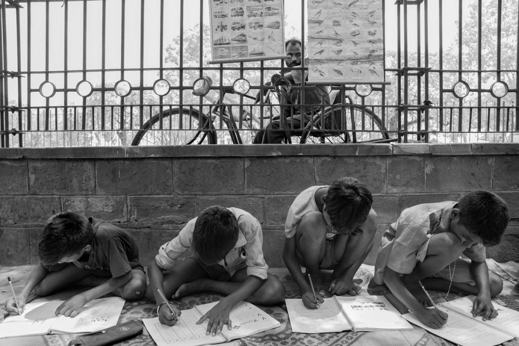 Extérieur noir et blanc. A Delhi. Quatre jeunes enfants sur des nattes posées sur un trottoir sont concentrés sur leurs tracaux d'écriture. Ils font leur devoir d'écolier dans la rue. En surplomb séparé par une grille, un homme les observent