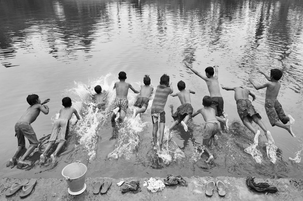 extérieur- photographie en noir et blanc montrant des jeunes garçons torses nus, en caleçons longs se jettant à l'eau. La photo les montre de dos. Avant de sauter/plonger, ils sont déposé leurs tongs et tee-shirts sur une margelle en pierre