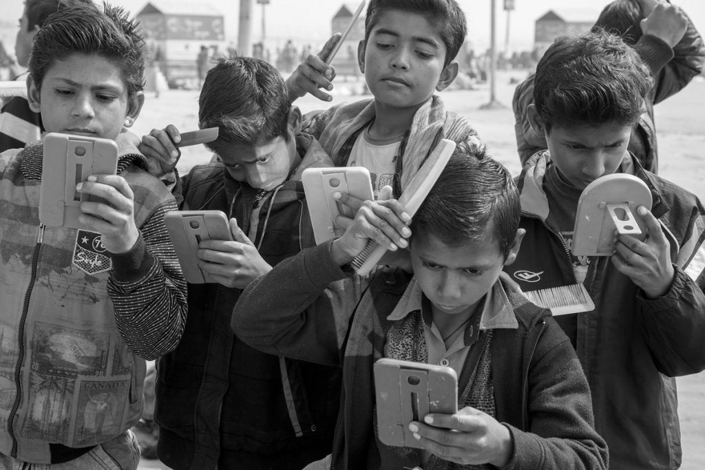 Extérieur noir et blanc. Six grands coquets, jeunes garçons lissent leurs cheveux en se regardant dans la vitre de leurs téléphone.