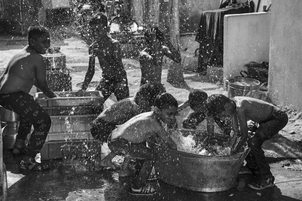 Extérieur noir et blanc. De jeuens garçons jouent à s'asperger penchés sur de grandes bassines dont ils extraient l'eau à deux mains. Des goutelettes en contre-jour semblent ruisseler du ciel.