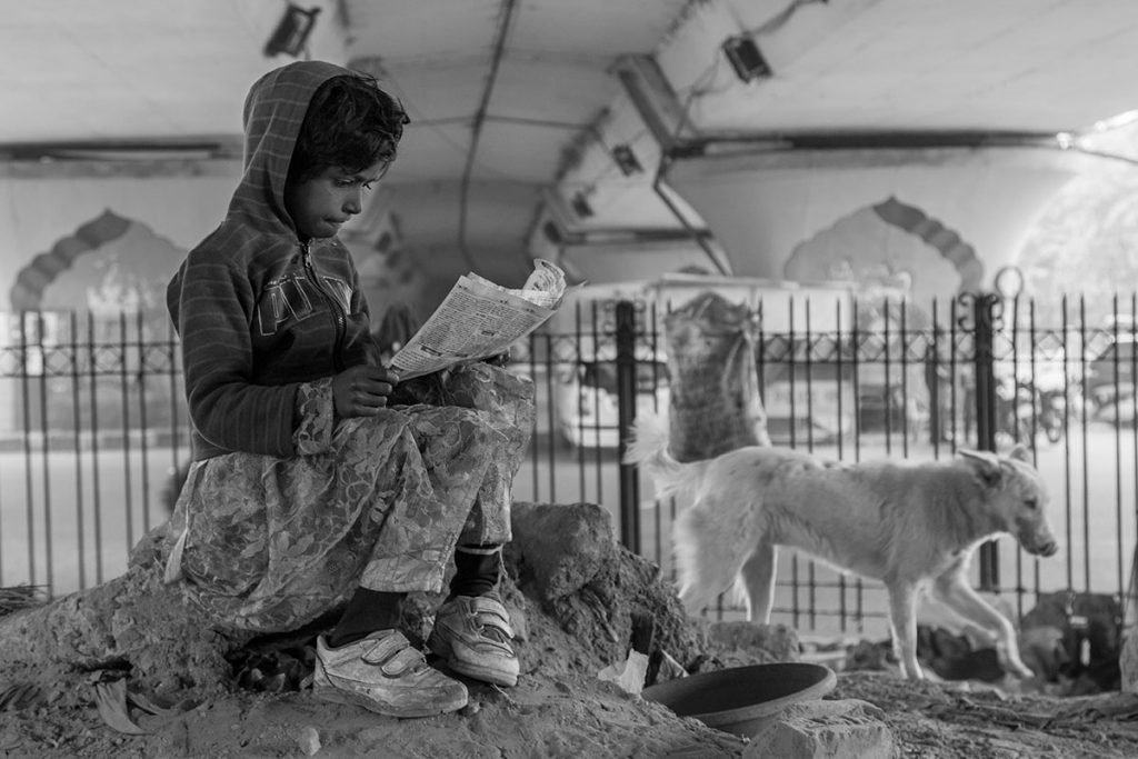 Exterieur noir et blanc. Dans Delhi. Ue jeune fille pauvrement vêtue assise au somment d'une petite butte de terre lit le journal. Un chien blanc passe à ses côtés. En arrière plan, une grille puis la route.
