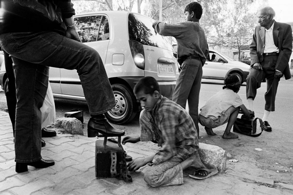 Exterieur noir et blanc. Dans Delhi. Deux jeunes garçons s'affairent au pied d'hommes bien habillés. Ils sont cireurs de chaussures. L'un porte des tongs, l'autre va nus-pieds.