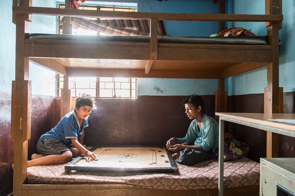 Intérieur - Maison de Salaam Baalak Trust. Dans un dortoir duquel on voit deux lits superposés. Ambiance léger contre jour. Deux enfant jouent à un grand jeu de plateau posé sur un lit.