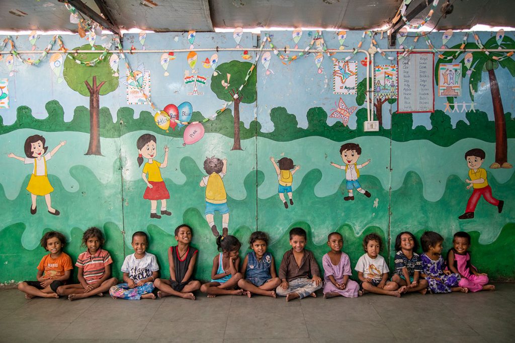 Lieu : maison de Salaam Baalak Trust. Salle de classe des petits. Devaun mur peint (représentat des enfants pratiquant des jeux de plein air dans un parc boisé) douze sont assis en tailleur adossés au mur, souriant et amusés