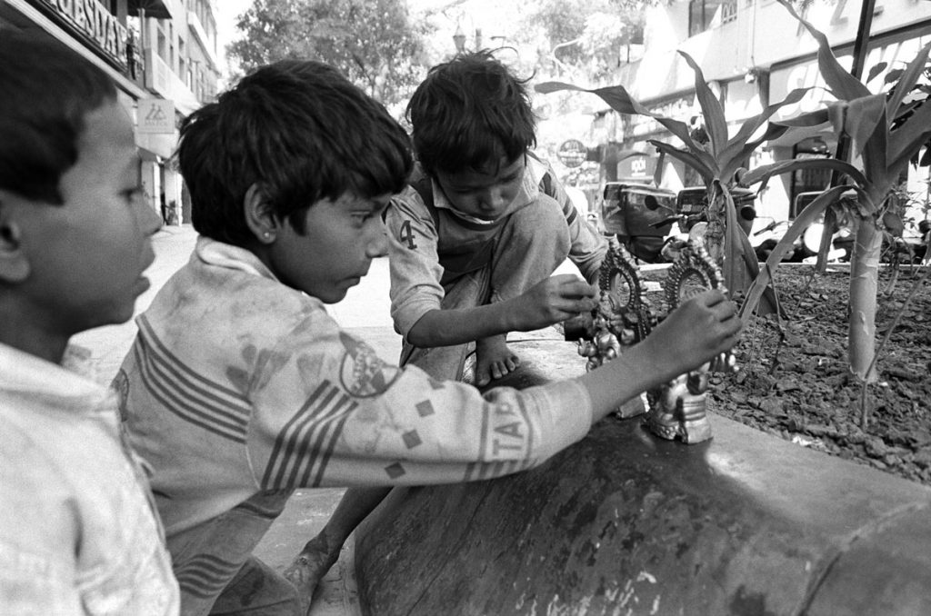 extérieur noir et blanc, dans les rues de Delhi. Trois enfants jouent avec des petites statues.