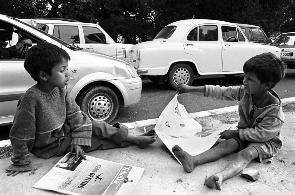 Extérieur boir et blanc. Sur un trottoir de New delhi, deux jeunes enfants assis lisent la presse.
