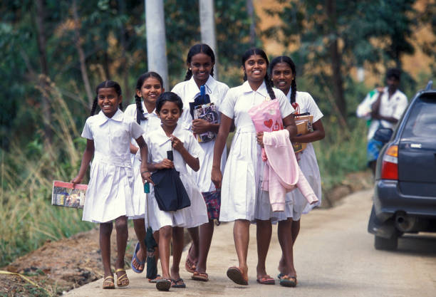 Un groupe de six jeunes filles âgées de 9 à 16 ans. Elles portent un uniforme jupe et chemiser blancs, elles sont joyeuses. Elles marchent au bord d'une route probablement en direction de l'école.