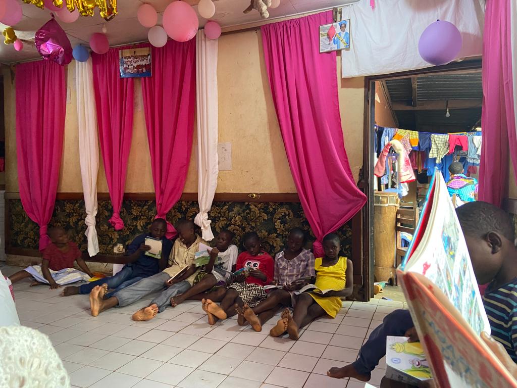 Plusieurs enfants pensionnaires de l'orphelinat de Ste Rica sont assis par terre les uns à côté des autres. Ils lisent chacun un livre. La pièce dans laquelle ils se trouvent est très colorée.