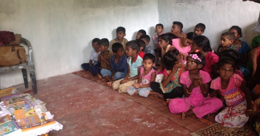 une vingtaine d'enfants assis par terre. un sol de brique. la pièce est modeste. les enfants attendent la distribution des livres.