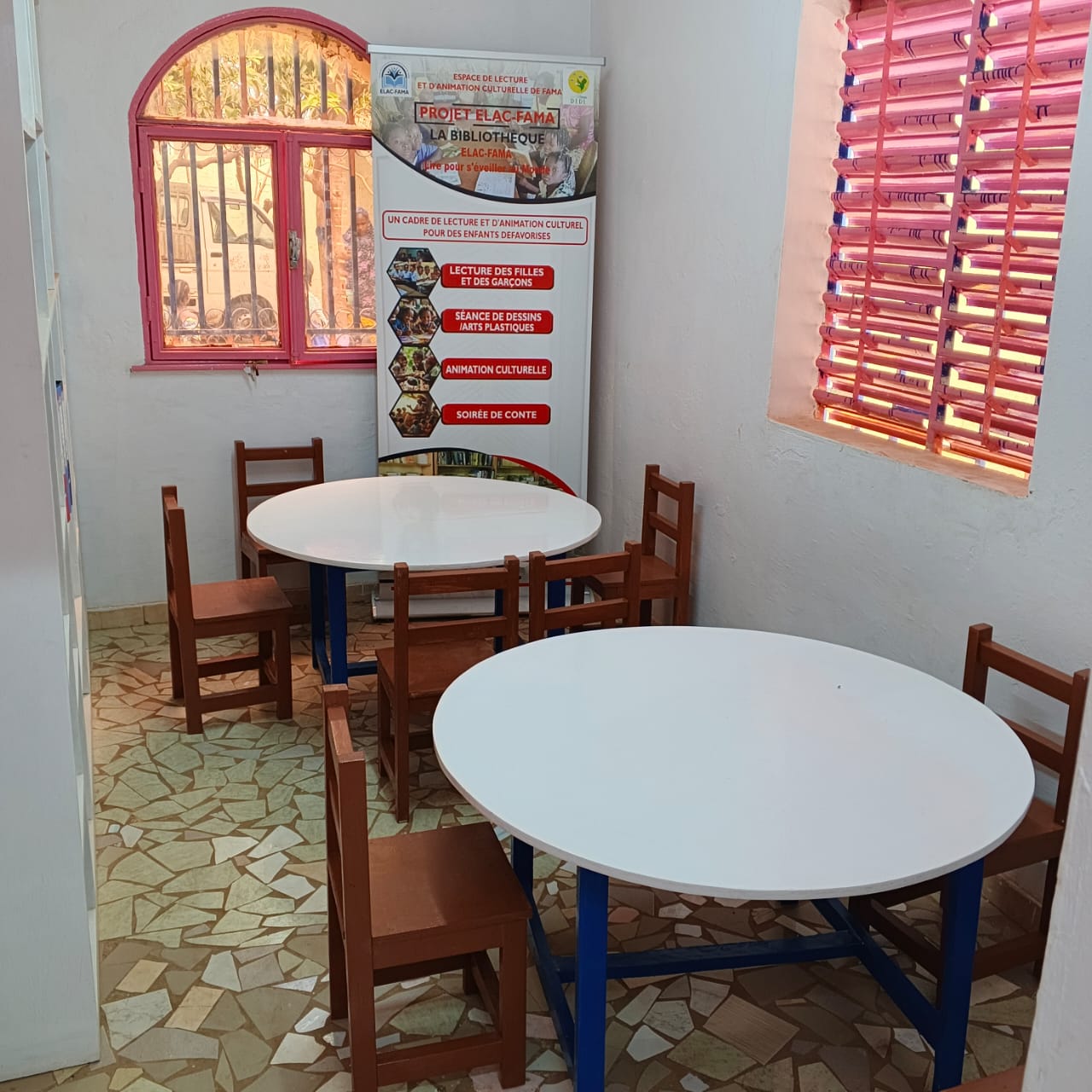 une petite pièce sol carrelé, deux fenêtres avec barreau, deux tables ronde avec des chaises.