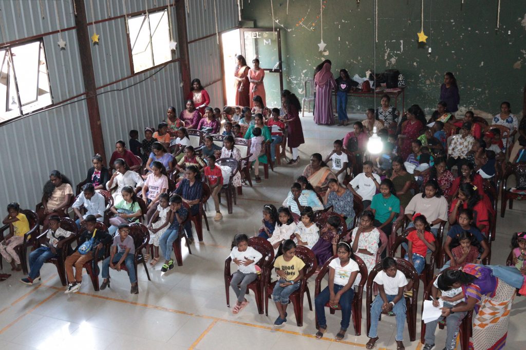une grande salle comme un hangar. Des dizaines d'enfants assis sur des chaises.